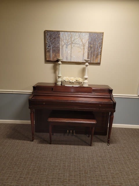 A wooden piano with a matching bench sits in a room, decorated with candles and a wall painting above it.