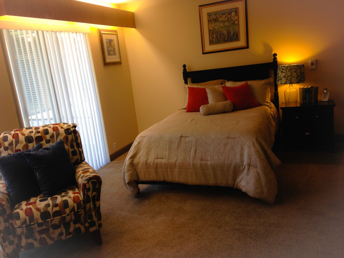 A cozy bedroom with a neatly made bed, accented by red and beige pillows, a patterned chair, and warm lighting.