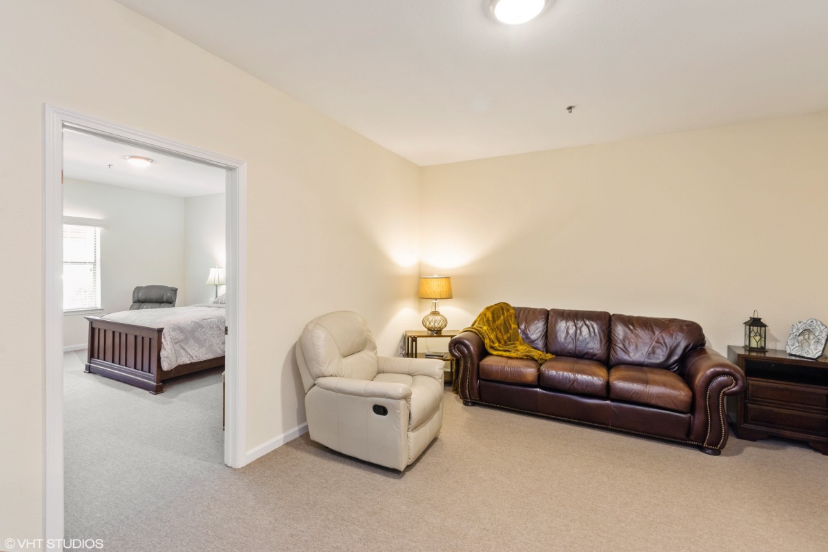 A cozy living room featuring a leather sofa, an armchair, and a lamp placed in the corner. A doorway leads to a bedroom.