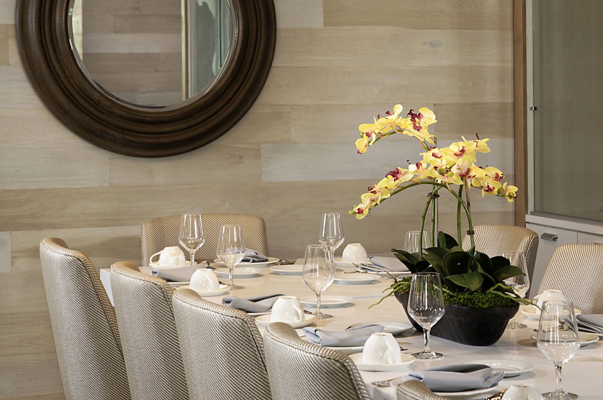 An elegantly set dining table with a yellow orchid centerpiece and wine glasses, surrounded by plush chairs and accentuated by a large round mirror on the wall.