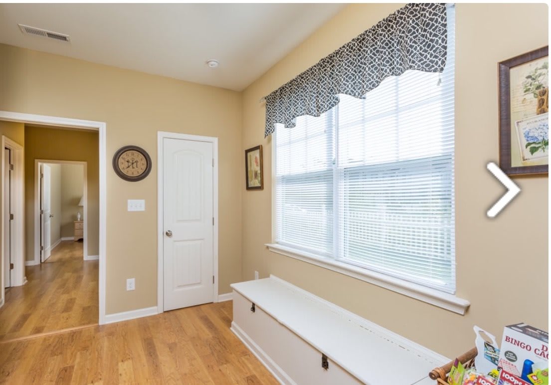 A bright hallway featuring a window with patterned curtains, a bench, and hardwood flooring.