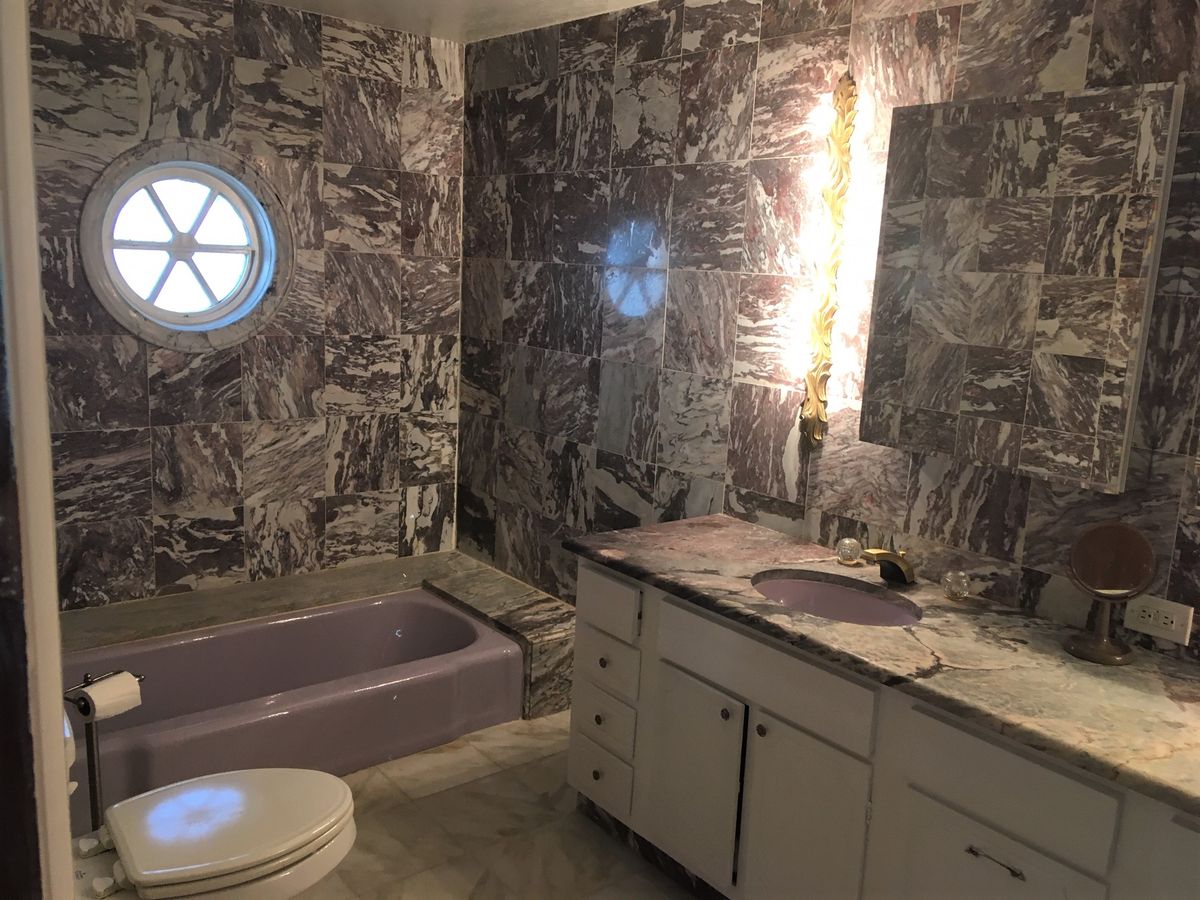 A bathroom featuring marble-patterned walls, a lavender bathtub, and a round window.