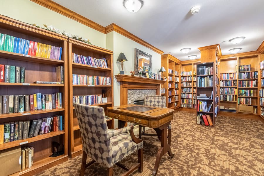 A cozy library with wooden bookshelves, a checkered chair, and a central wooden table in front of a fireplace.