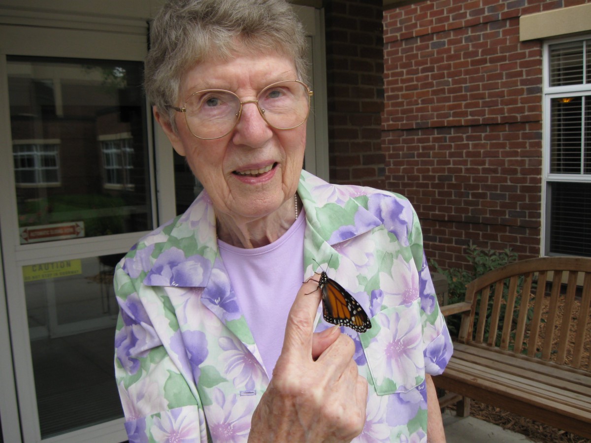 An elderly woman smiling while holding a butterfly on her hand.