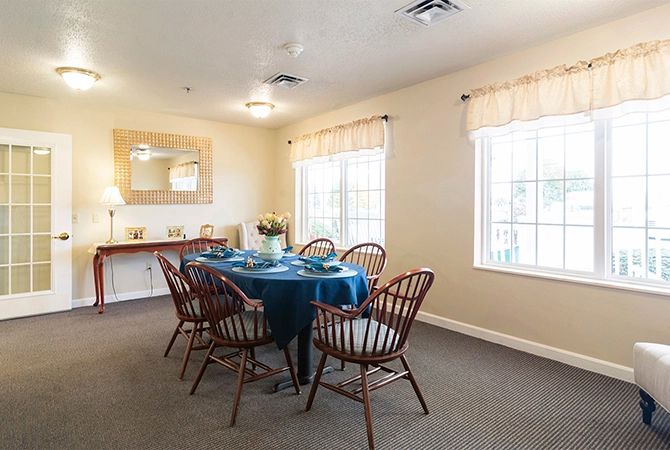 A cozy dining room with a set table, wooden chairs, and natural light from large windows.
