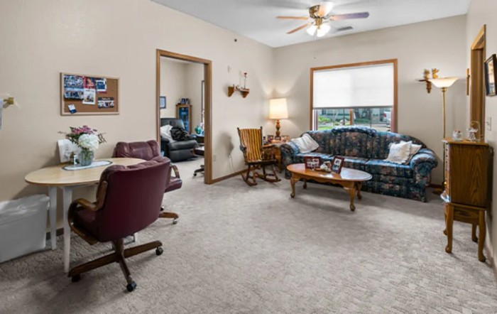 A cozy living room with a sofa, desk, and lamp, featuring carpeted flooring and a window.