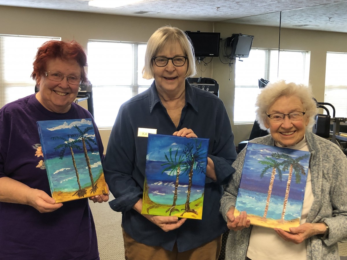 A group of three individuals proudly displaying their colorful paintings of beach scenes with palm trees.