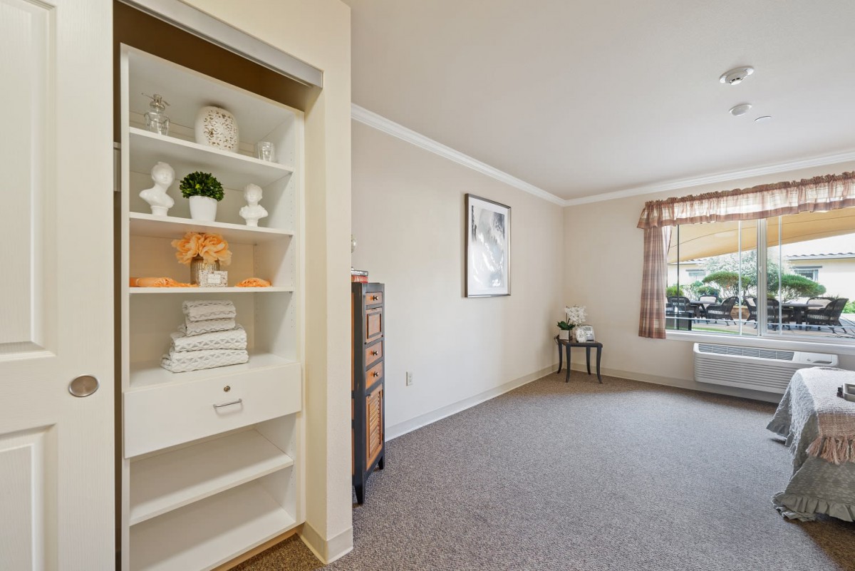 A cozy living room with a built-in shelf and carpet flooring, featuring natural light from a window with checkered curtains.