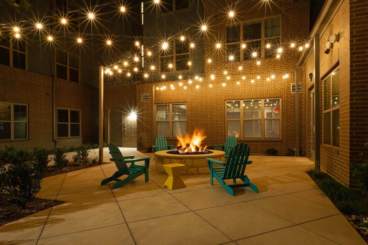 A cozy outdoor patio with string lights and a central fire pit surrounded by colorful chairs.