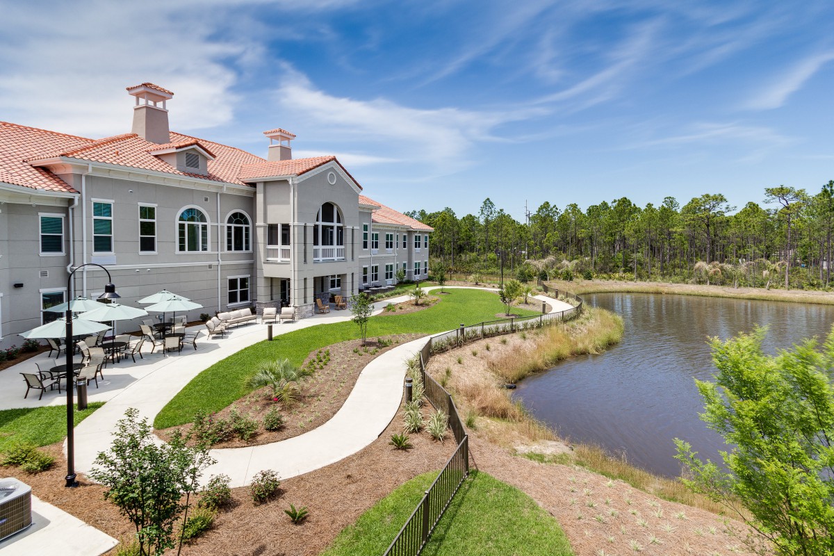 A large, elegant building with a red roof by a scenic lake, surrounded by landscaped pathways and greenery under a blue sky.