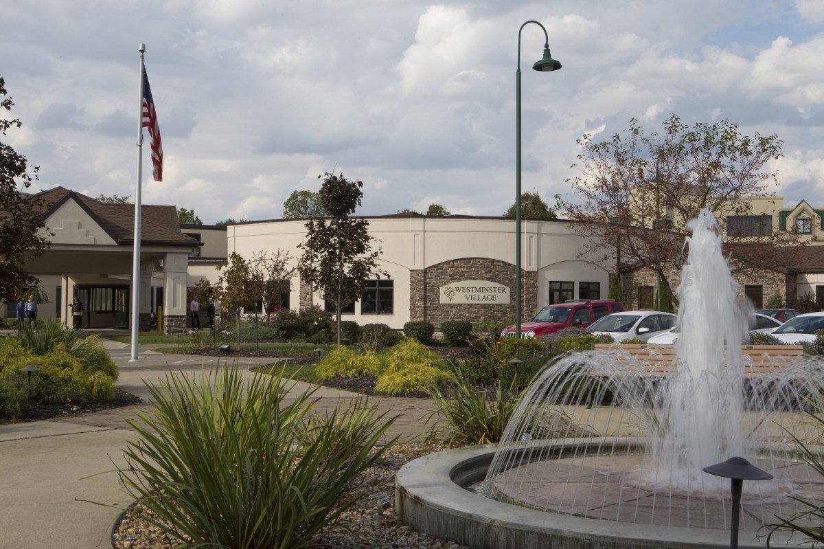 A building with a fountain in the foreground, accompanied by a flag and a parking area under a cloudy sky.