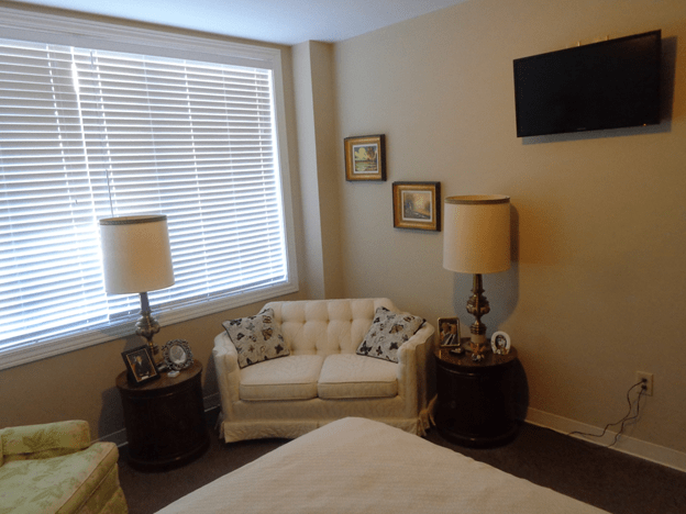 A cozy bedroom with classic decor, featuring a seating area with lamps and a window with blinds.