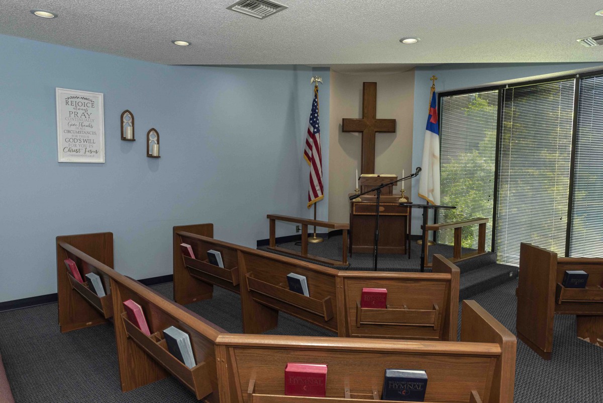 Interior of a small chapel with wooden pews, a cross, and flags at the front.