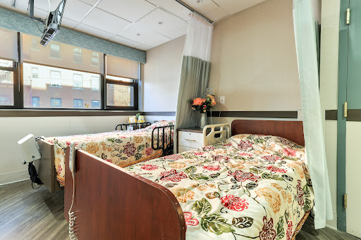A well-lit hospital patient room featuring two beds with floral bedding and a window view.
