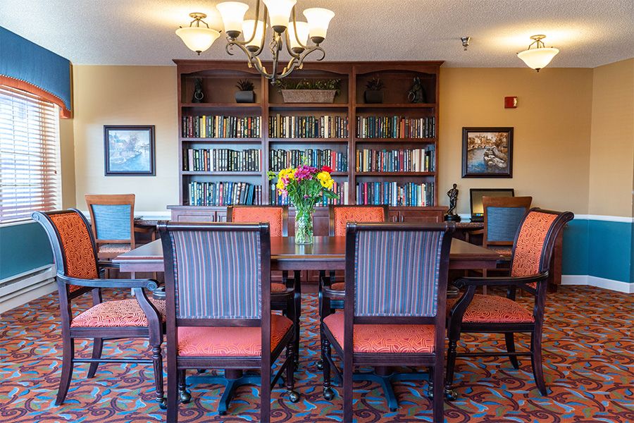 A cozy library room with a large wooden table, surrounded by chairs and a bookshelf filled with books. A vase of colorful flowers adorns the center of the table, and a chandelier hangs from the ceiling.