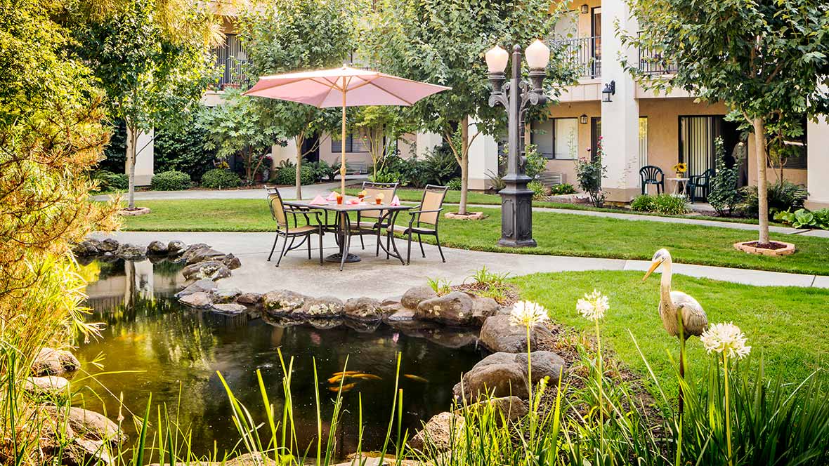 A tranquil garden setting with patio furniture under an umbrella, surrounded by greenery and a small pond.