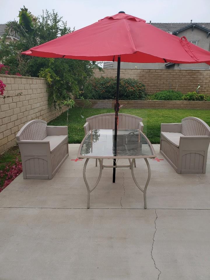 An outdoor patio setup with a red umbrella, glass table, and two chairs in a garden setting.