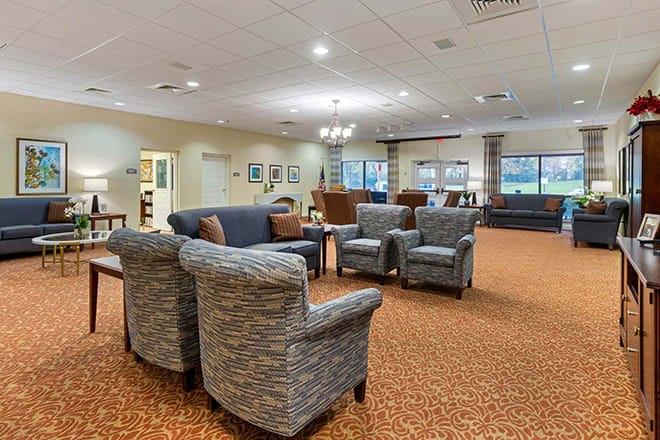 A cozy living room with comfortable seating, including sofas and armchairs, carpeted flooring, and ceiling lights.