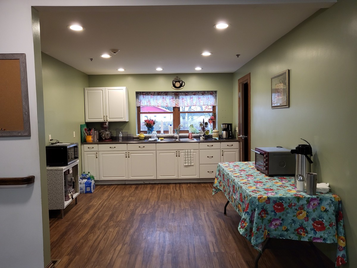 A cozy kitchen with green walls, featuring cabinets, a wooden floor, and a table covered with a floral tablecloth.