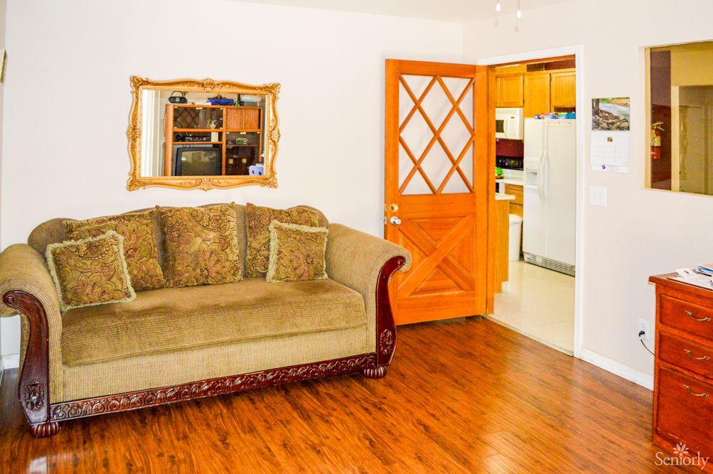 A cozy living room with a plush sofa, elegant mirror, and a view into the kitchen through an open wooden door.
