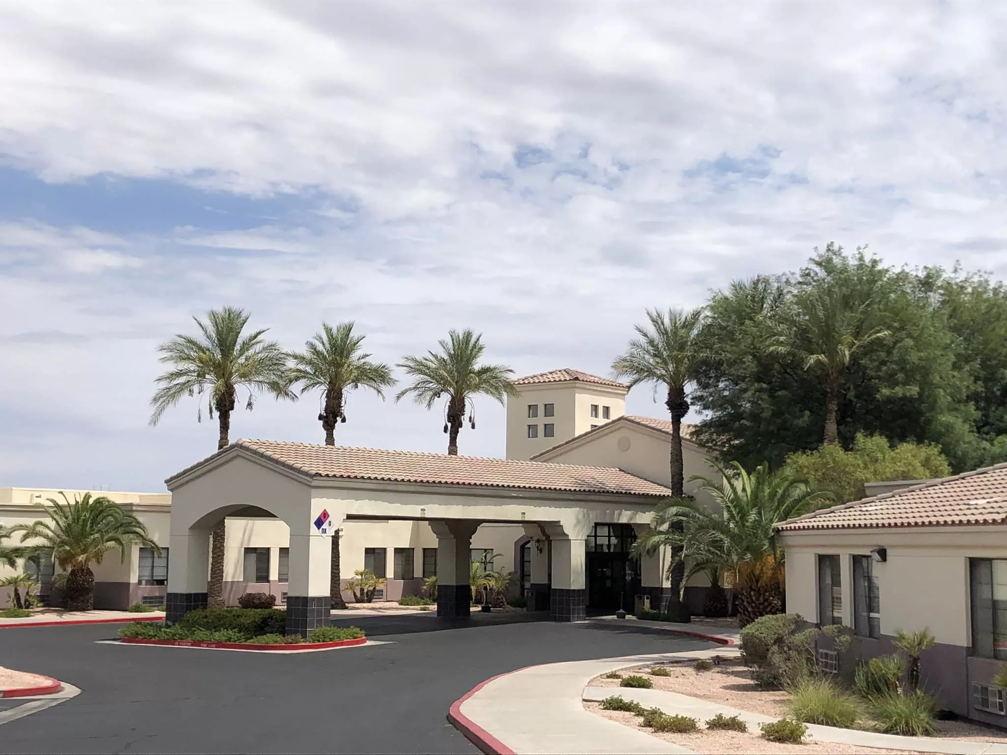A building entrance with palm trees lining the driveway under a cloudy sky.