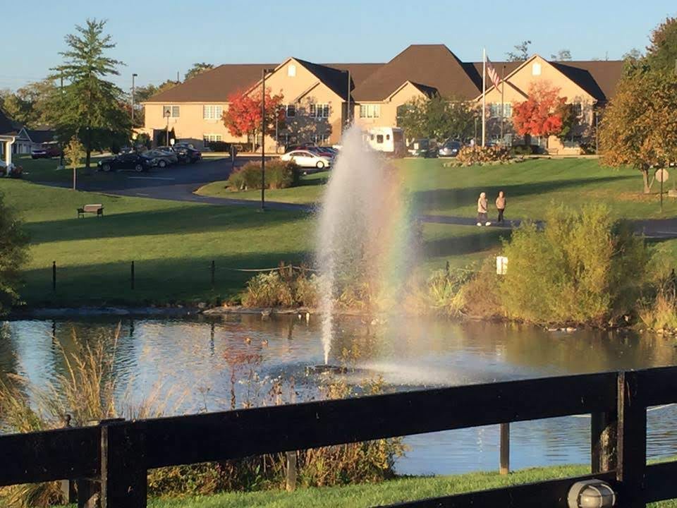 A tranquil park scene featuring a fountain creating a rainbow over a pond, with autumn trees and houses in the background.