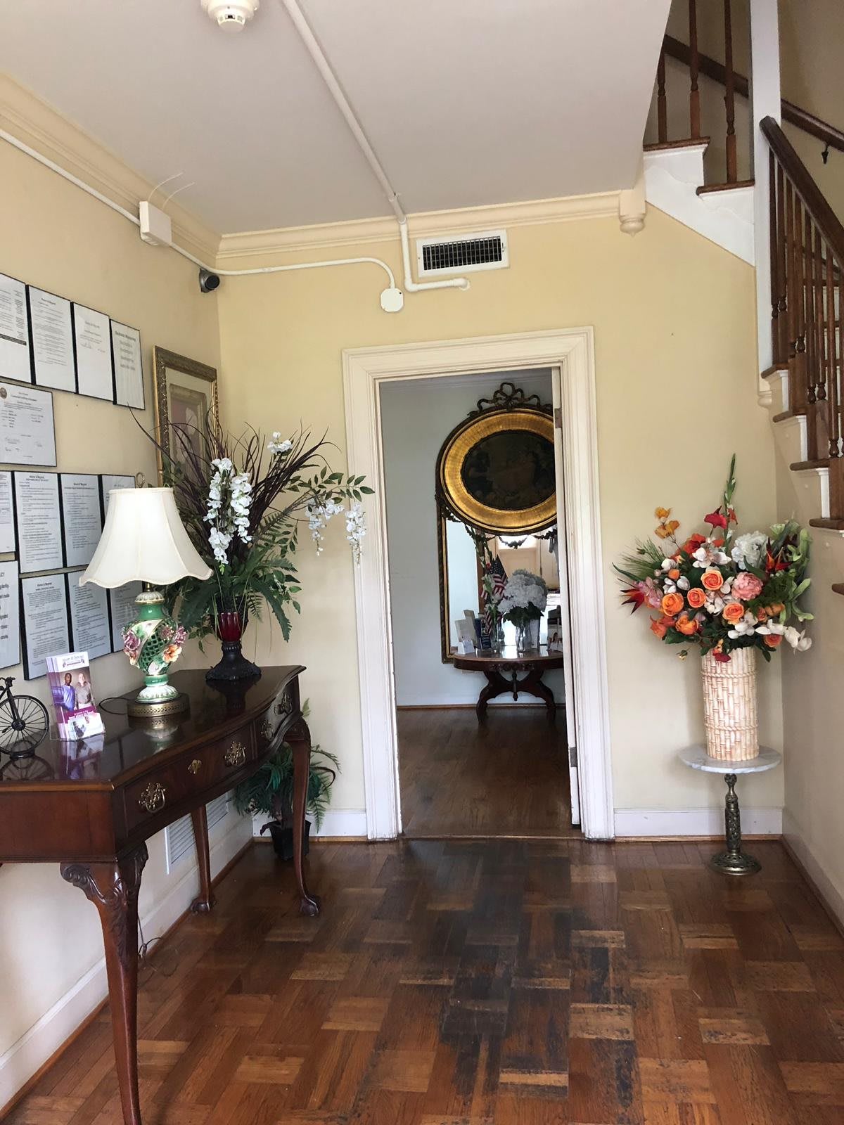 A well-decorated hallway featuring a table with a lamp and plant, framed certificates on the wall, and a staircase leading upwards. A large mirror is visible through the doorway along with colorful floral arrangements.