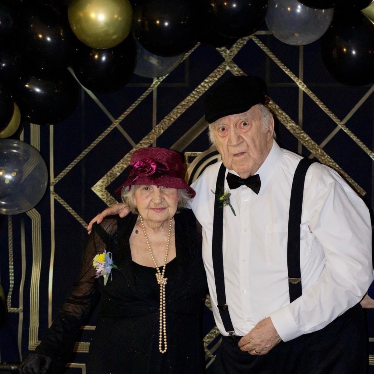 An elderly couple dressed in vintage black-tie attire pose at a themed party with decorative balloons.