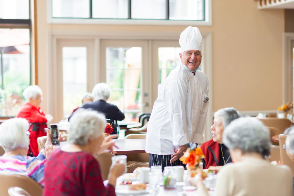 A chef interacts with seniors in a dining area, creating a welcoming atmosphere.