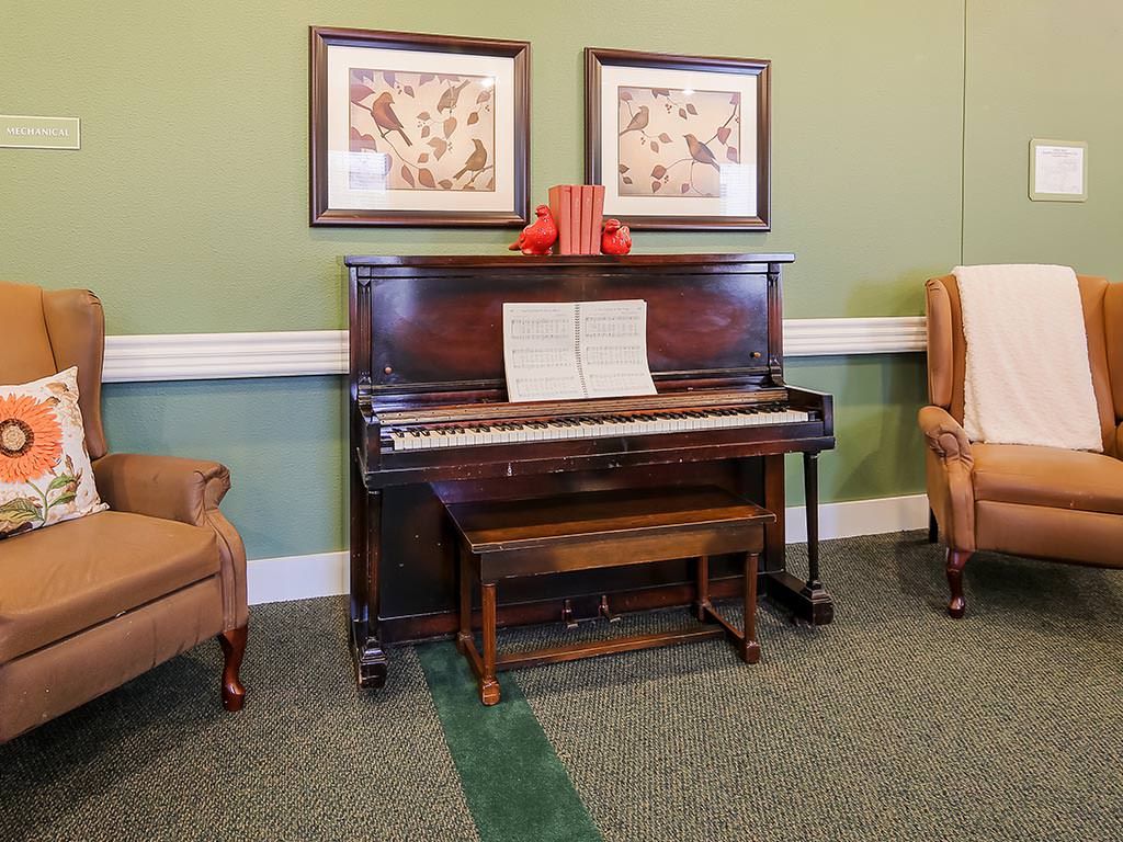 A cozy room featuring a vintage upright piano with sheet music and two armchairs, adorned with framed artwork above.