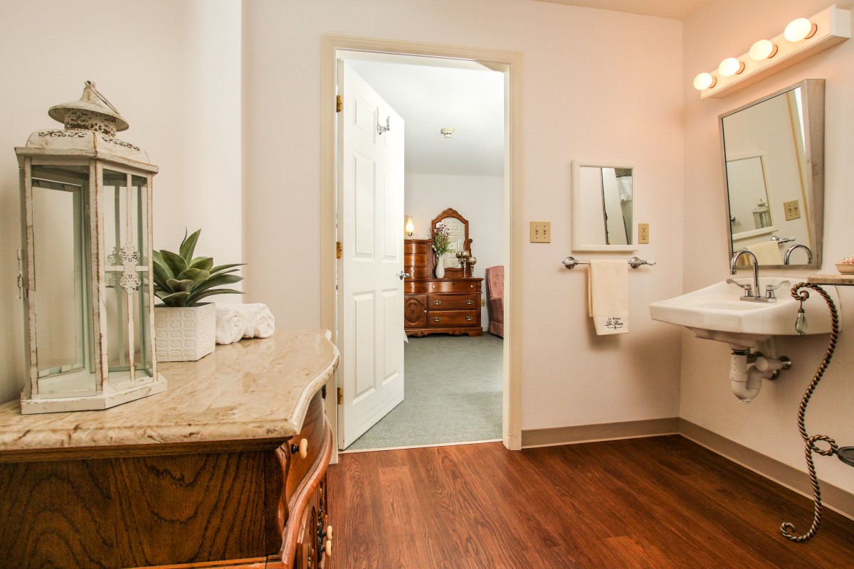 A view of a bathroom with wooden flooring, featuring a sink, mirror, towel rack, and plant, leading into a bedroom with furniture.