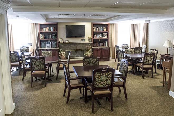 A cozy living room with tables and chairs, a television mounted above a fireplace, and bookshelves lining the walls.