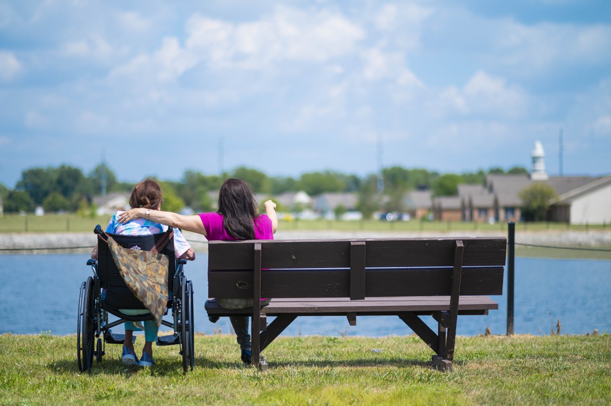 Two people enjoying a peaceful moment by the lake, one sitting in a wheelchair and the other on a bench.