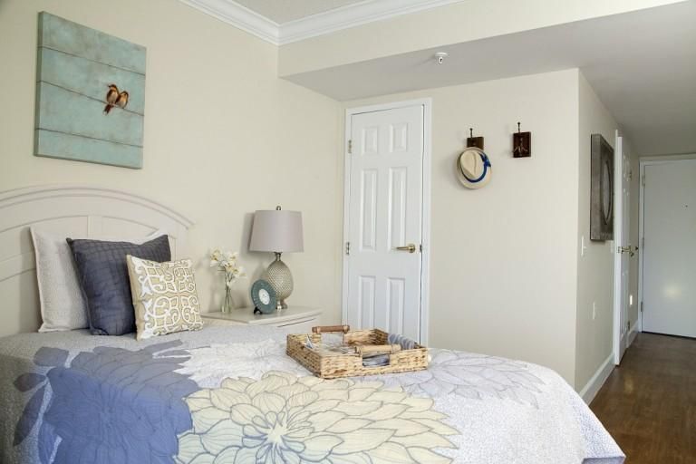 A cozy bedroom with light decor, featuring a bed with floral bedding, a bedside lamp, and wall decorations including artwork and a clock.