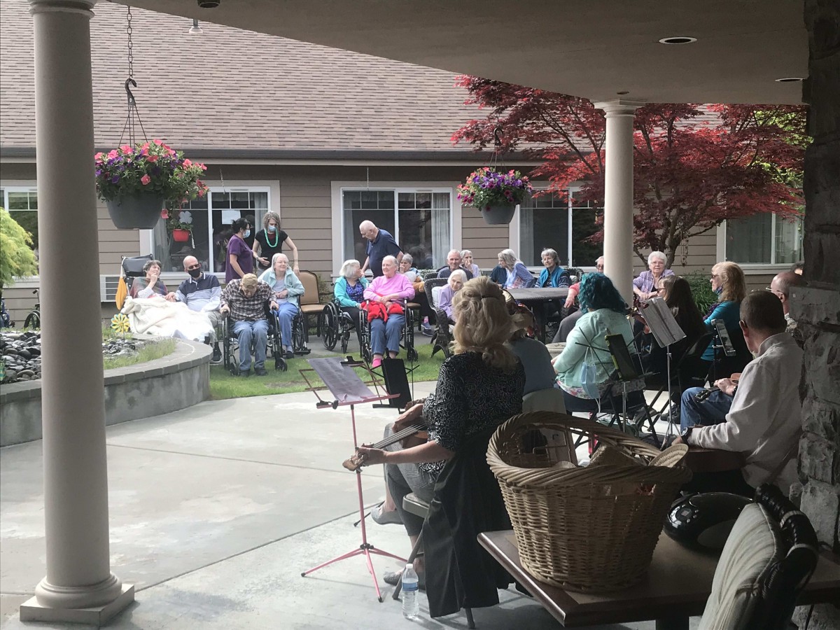 A group of musicians performs in front of an audience seated outdoors in a garden setting.