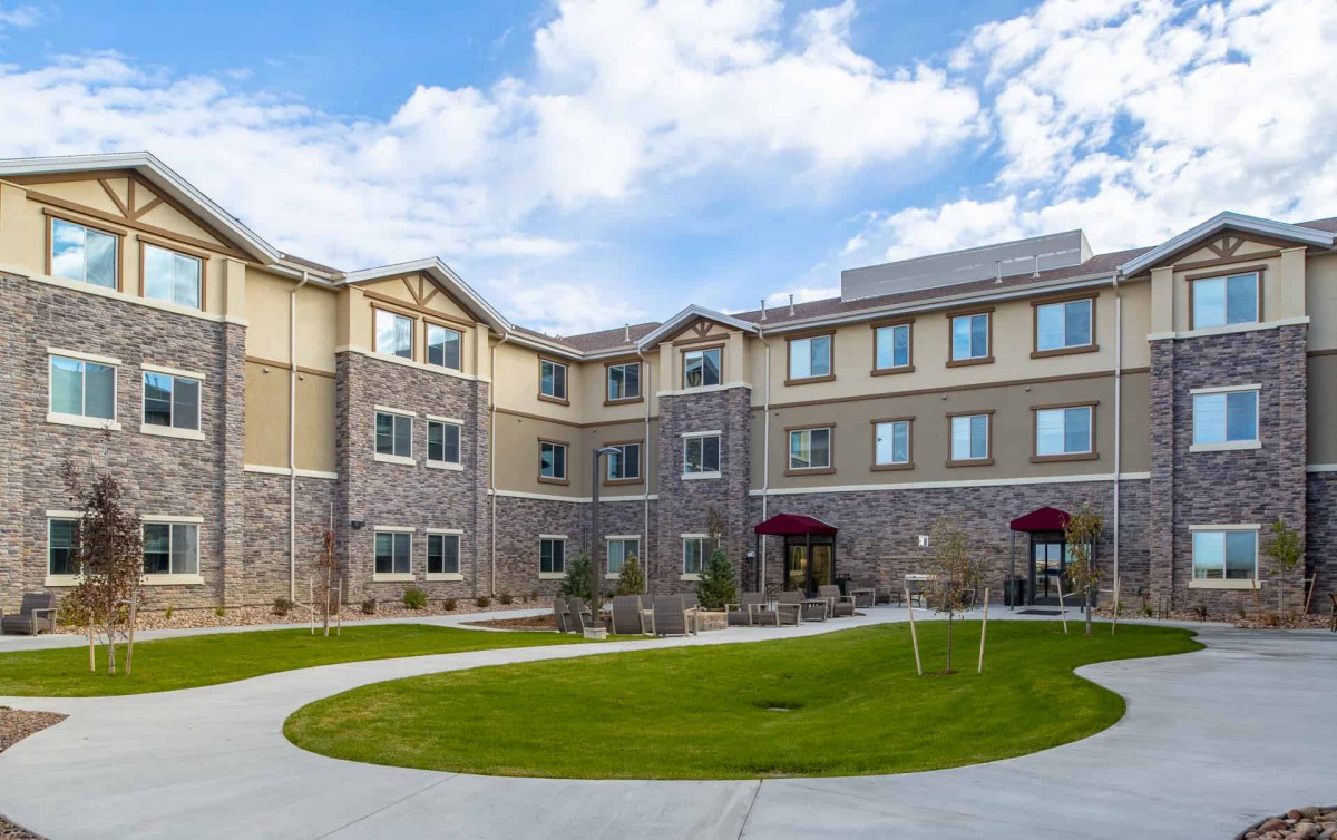 A view of a multi-story residential building with a stone and stucco exterior, surrounded by a well-maintained lawn under a partly cloudy sky.