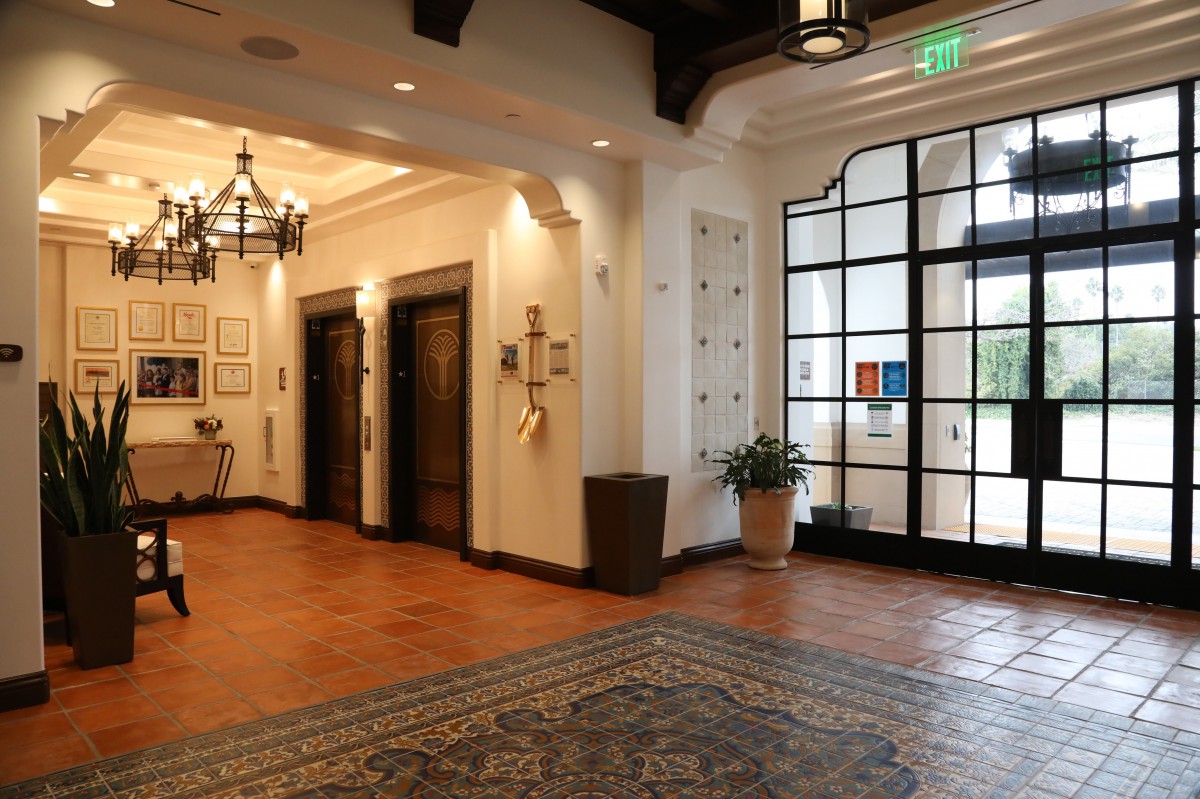 A lobby area with tile flooring, featuring two elevators and a glass entrance door, elegantly lit by a chandelier.