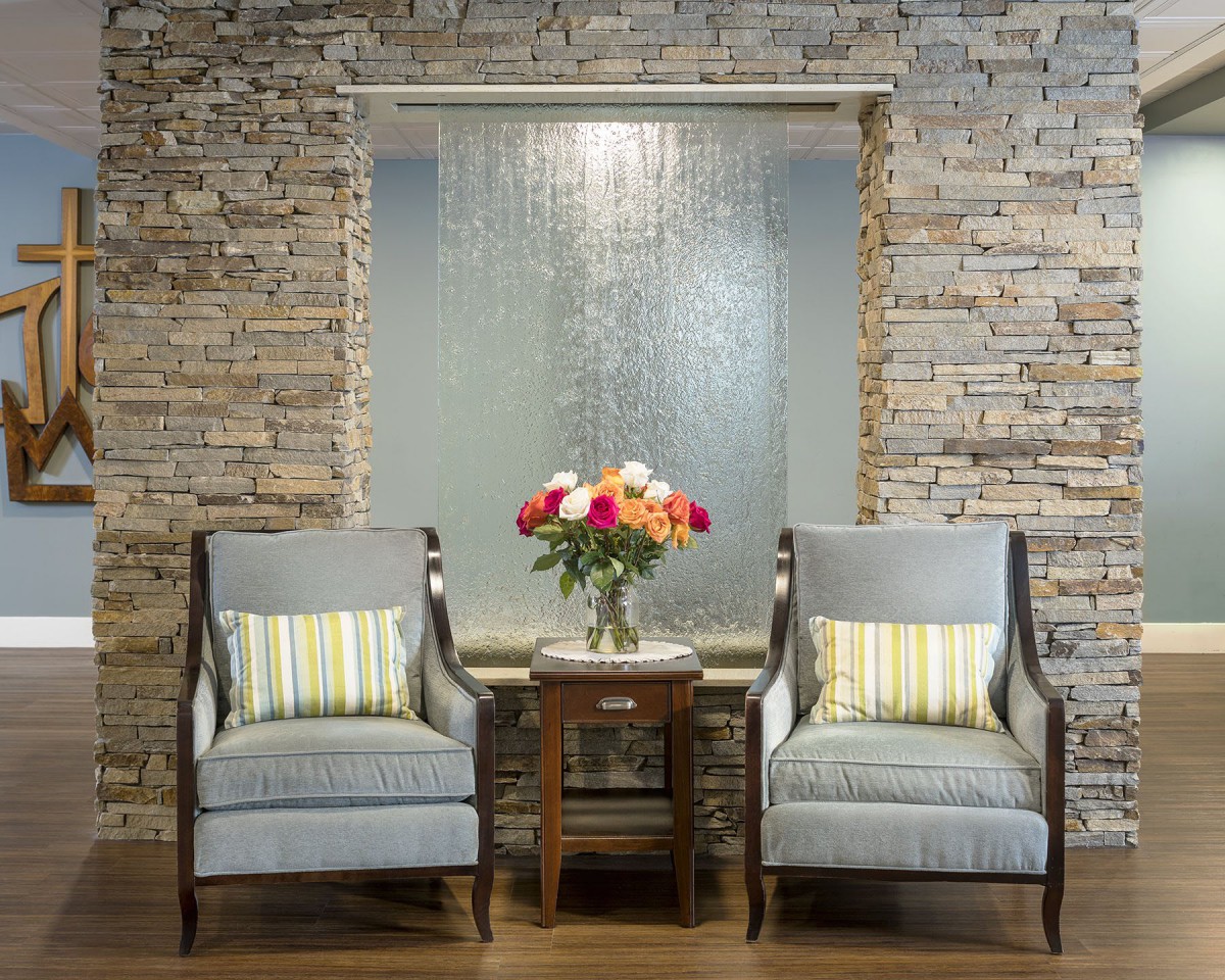 A cozy seating area featuring two chairs with striped pillows, a table with a vase of flowers, set against a brick wall and water feature.