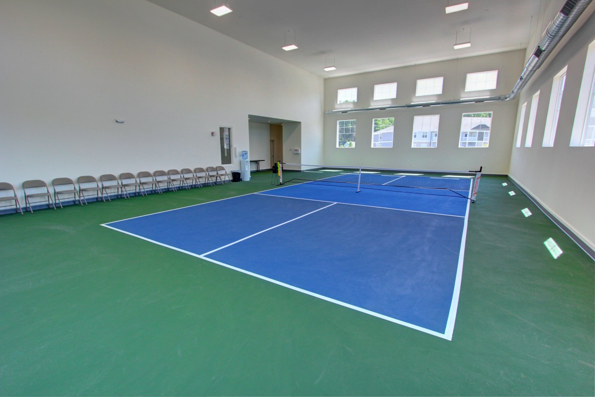 An indoor sports facility featuring a blue tennis court with a green floor, surrounded by windows and lined with chairs.