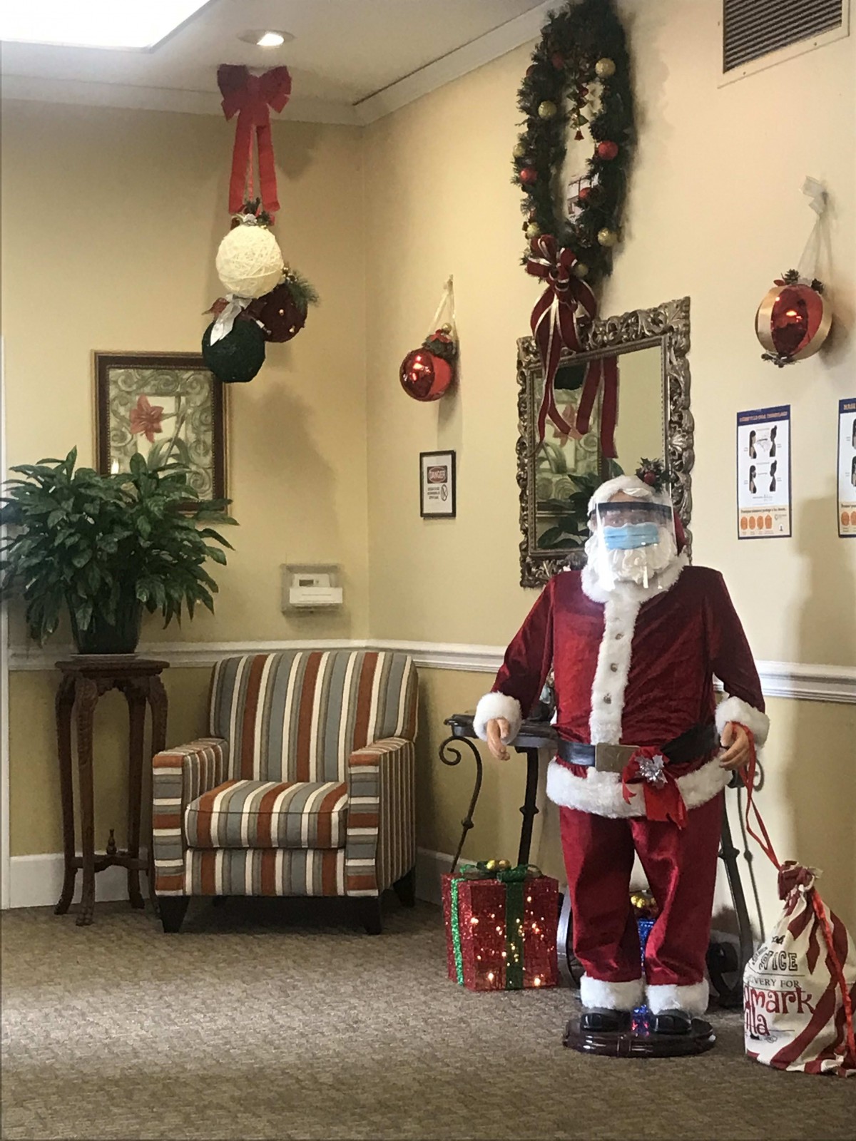 A Santa Claus figure wearing a protective mask stands in a festively decorated room.