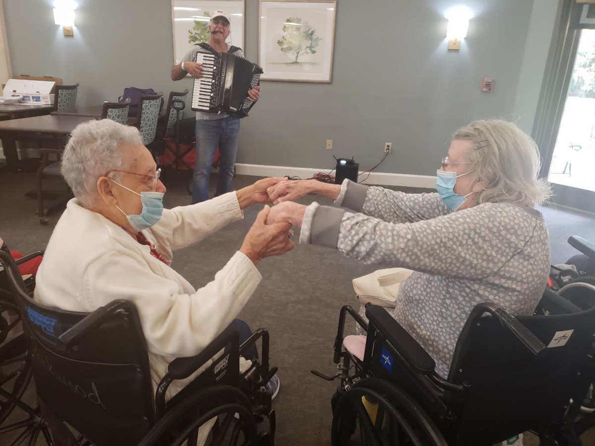 Two elderly individuals in wheelchairs holding hands and wearing masks in a care facility.
