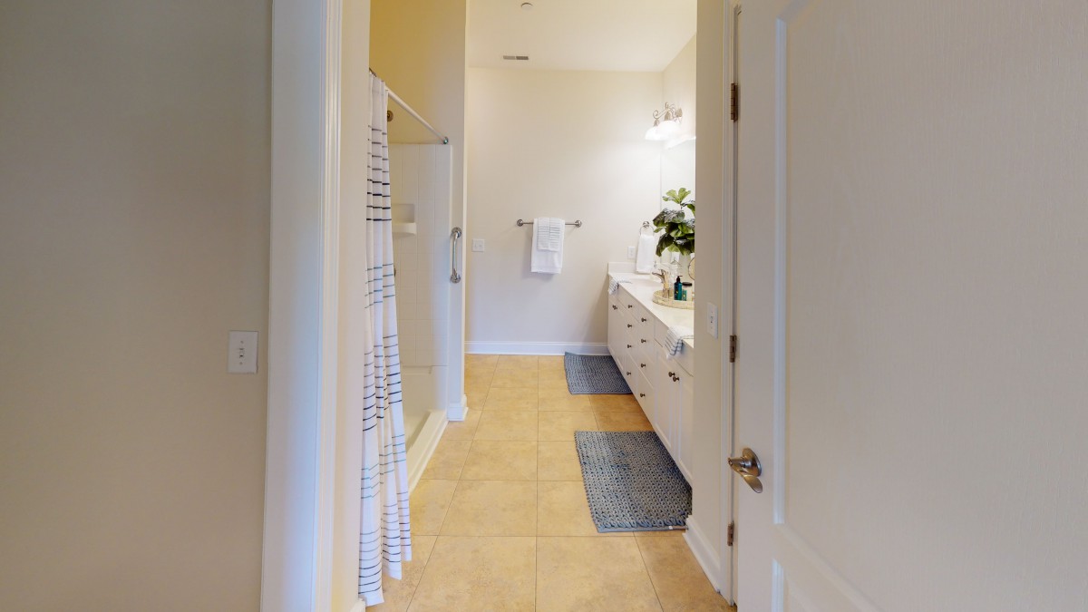 A well-lit bathroom featuring a shower, double sink vanity with mirrors, and floor mats.