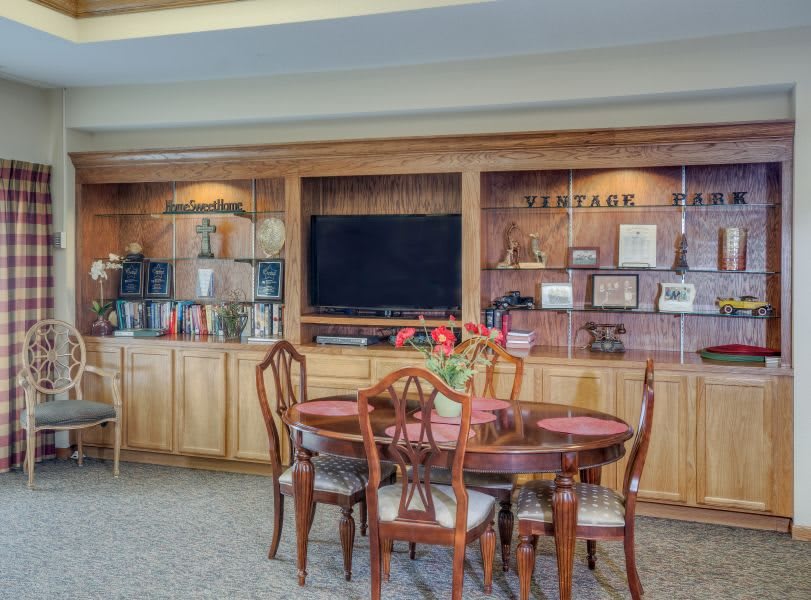 A cozy living room with wooden furniture, featuring a dining table and a television on wooden shelves adorned with decorative items.