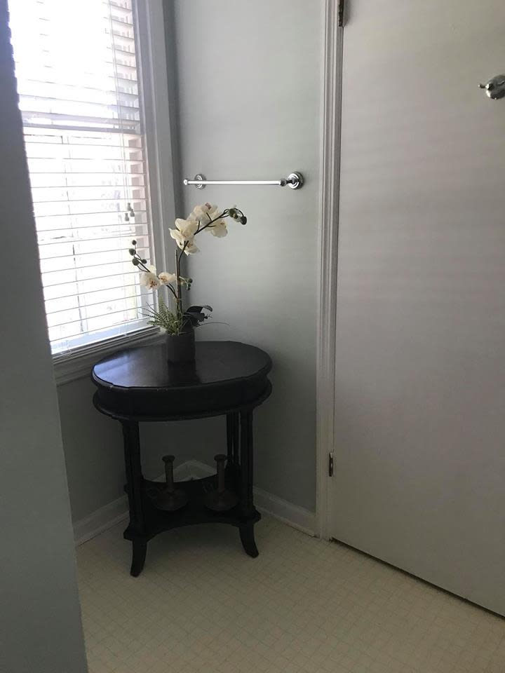 A dark round table with a vase of flowers in a bright corner near a window and door.
