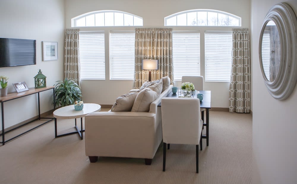 A cozy living room with a neutral color palette, featuring a sofa, coffee table, dining table, and large windows with curtains.
