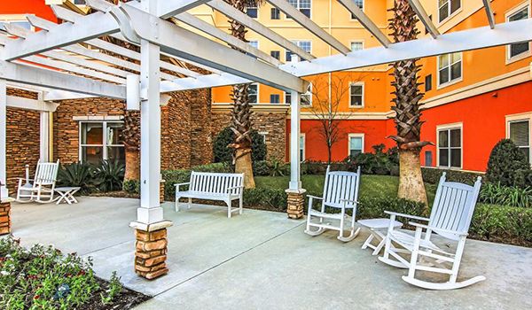 A cozy outdoor seating area with benches and rocking chairs under a pergola, surrounded by lush landscaping and palm trees.
