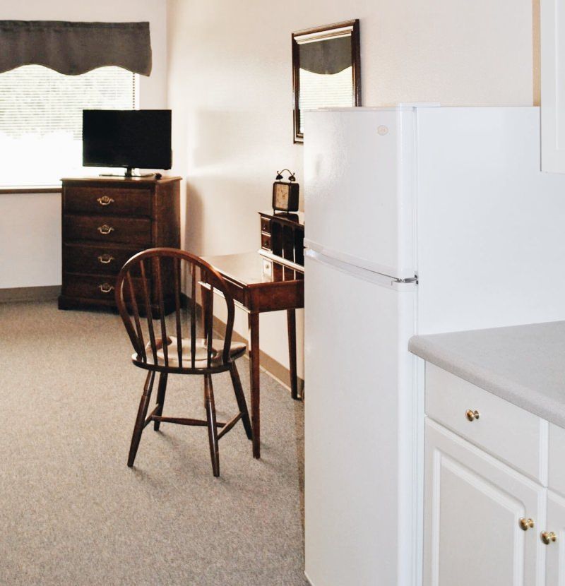 A cozy apartment interior with a wooden desk and chair, a dresser with a television, and a refrigerator.