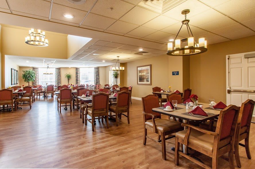 A spacious dining room with elegant chandeliers, neatly arranged tables and chairs, and a warm wooden floor.