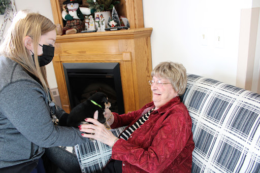 A joyful moment shared between a caregiver and an elderly woman on a cozy couch, with a playful dog nearby.