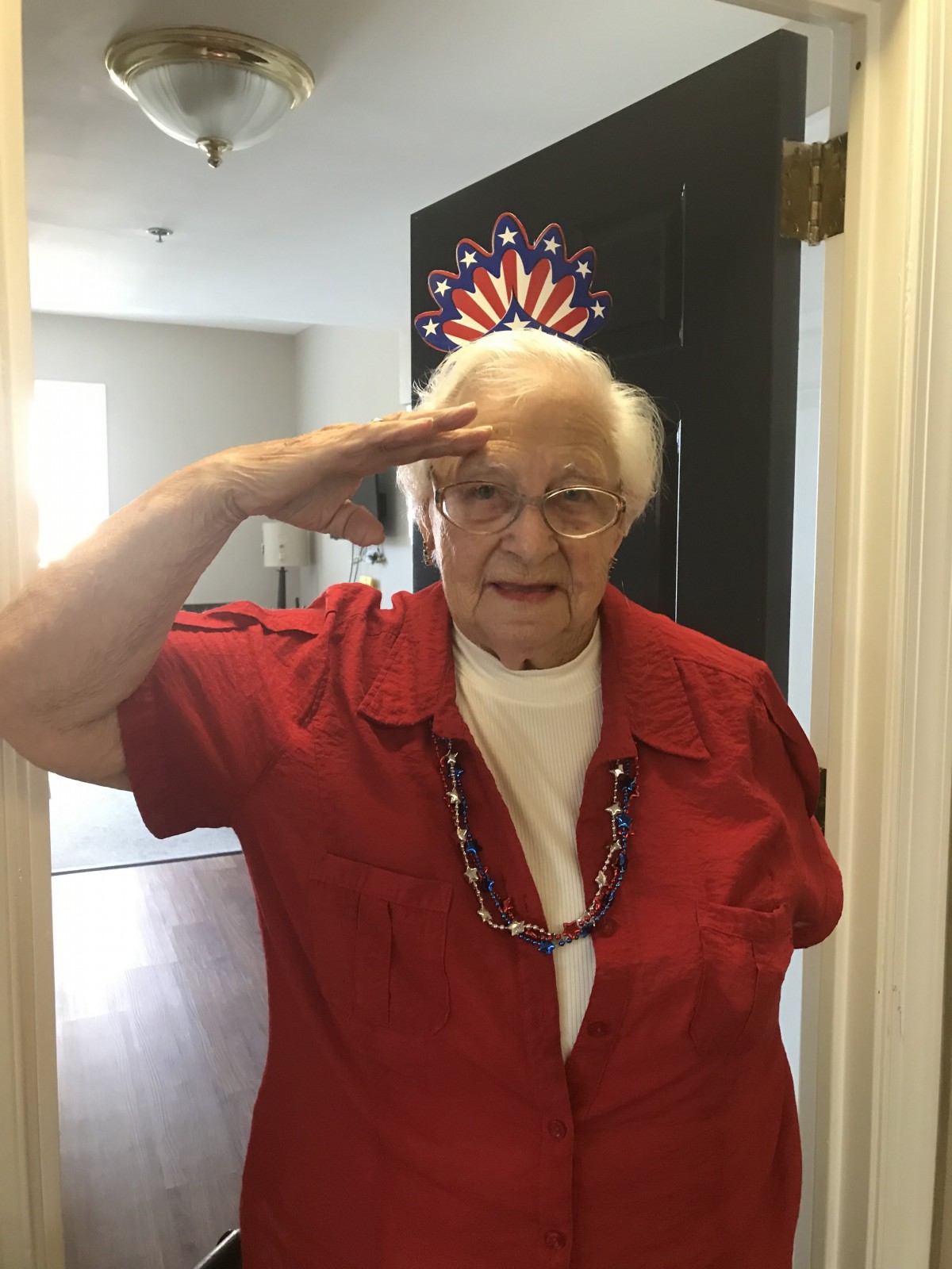 An elderly woman in a red shirt giving a salute indoors.